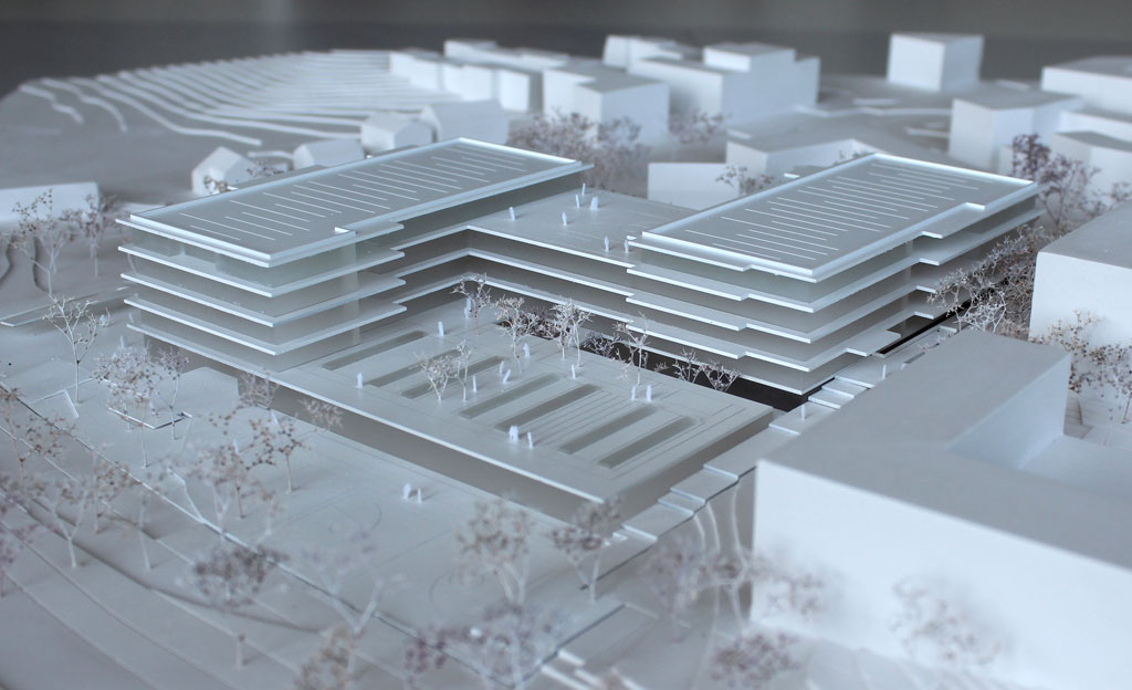 Foto von einem weißen Architekturmodell mit einfachen Kuben für die Umgebungsgebäude und dem detaillierter ausgeführten Schulneubau. Dieser besteht aus zwei parallelen länglichen Riegeln, die versetzt zu einander angeordnet sind und die mit einem senkrecht dazu verlaufenden schmäleren Querbau verbunden sind. Die Längsriegel bestehen jeweils aus 5 Geschossen, der Querriegel aus vier Geschossen. Jedes Geschoss ist aus mattiertem Plexiglas gefertigt, die auskragenden Böden und Decken aus weißem Kunststoff. Im Erdgeschoss gibt es eine Sporthalle, deren Dach begehbar und als Aufenthaltsfläche nutzbar ist. Die Bäume sind aus weiß lackierten getrockneten Pflanzen aufgeklebt.
