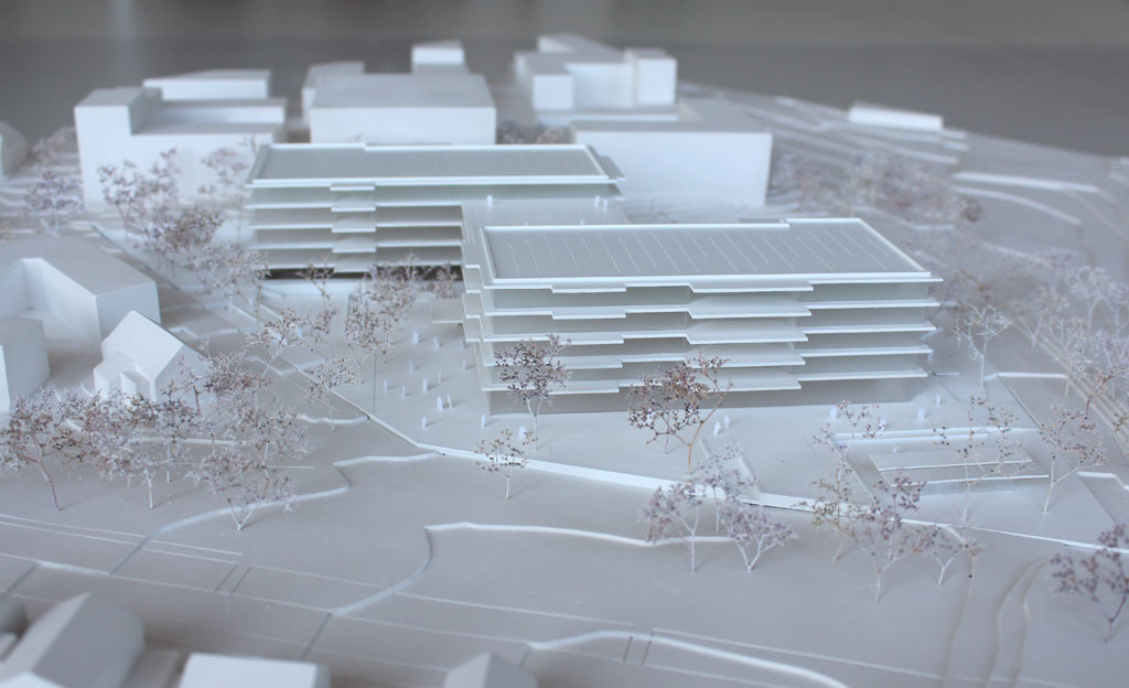 Foto von einem weißen Architekturmodell mit einfachen Kuben für die Umgebungsgebäude und dem detaillierter ausgeführten Schulneubau. Dieser besteht aus zwei parallelen länglichen Riegeln, die versetzt zu einander angeordnet sind und die mit einem senkrecht dazu verlaufenden schmäleren Querbau verbunden sind. Die Längsriegel bestehen jeweils aus 5 Geschossen, der Querriegel aus vier Geschossen. Jedes Geschoss ist aus mattiertem Plexiglas gefertigt, die auskragenden Böden und Decken aus weißem Kunststoff. Im Erdgeschoss gibt es eine Sporthalle, deren Dach begehbar und als Aufenthaltsfläche nutzbar ist. Die Bäume sind aus weiß lackierten getrockneten Pflanzen aufgeklebt.