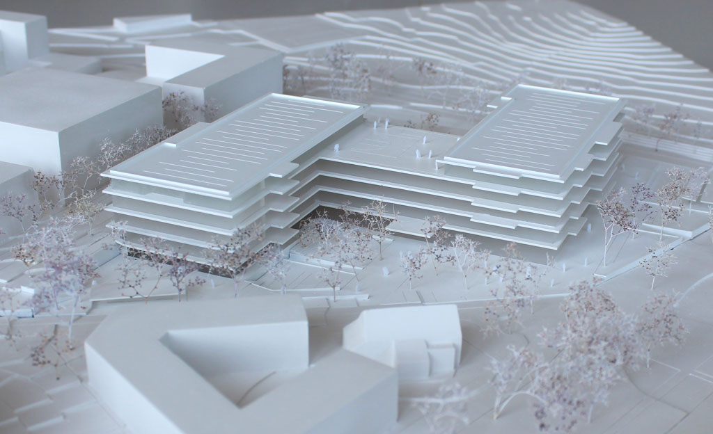 Foto von einem weißen Architekturmodell mit einfachen Kuben für die Umgebungsgebäude und dem detaillierter ausgeführten Schulneubau. Dieser besteht aus zwei parallelen länglichen Riegeln, die versetzt zu einander angeordnet sind und die mit einem senkrecht dazu verlaufenden schmäleren Querbau verbunden sind. Die Längsriegel bestehen jeweils aus 5 Geschossen, der Querriegel aus vier Geschossen. Jedes Geschoss ist aus mattiertem Plexiglas gefertigt, die auskragenden Böden und Decken aus weißem Kunststoff. Im Erdgeschoss gibt es eine Sporthalle, deren Dach begehbar und als Aufenthaltsfläche nutzbar ist. Die Bäume sind aus weiß lackierten getrockneten Pflanzen aufgeklebt.