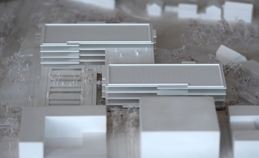 Foto von einem weißen Architekturmodell mit einfachen Kuben für die Umgebungsgebäude und dem detaillierter ausgeführten Schulneubau. Dieser besteht aus zwei parallelen länglichen Riegeln, die versetzt zu einander angeordnet sind und die mit einem senkrecht dazu verlaufenden schmäleren Querbau verbunden sind. Die Längsriegel bestehen jeweils aus 5 Geschossen, der Querriegel aus vier Geschossen. Jedes Geschoss ist aus mattiertem Plexiglas gefertigt, die auskragenden Böden und Decken aus weißem Kunststoff. Im Erdgeschoss gibt es eine Sporthalle, deren Dach begehbar und als Aufenthaltsfläche nutzbar ist. Die Bäume sind aus weiß lackierten getrockneten Pflanzen aufgeklebt.
