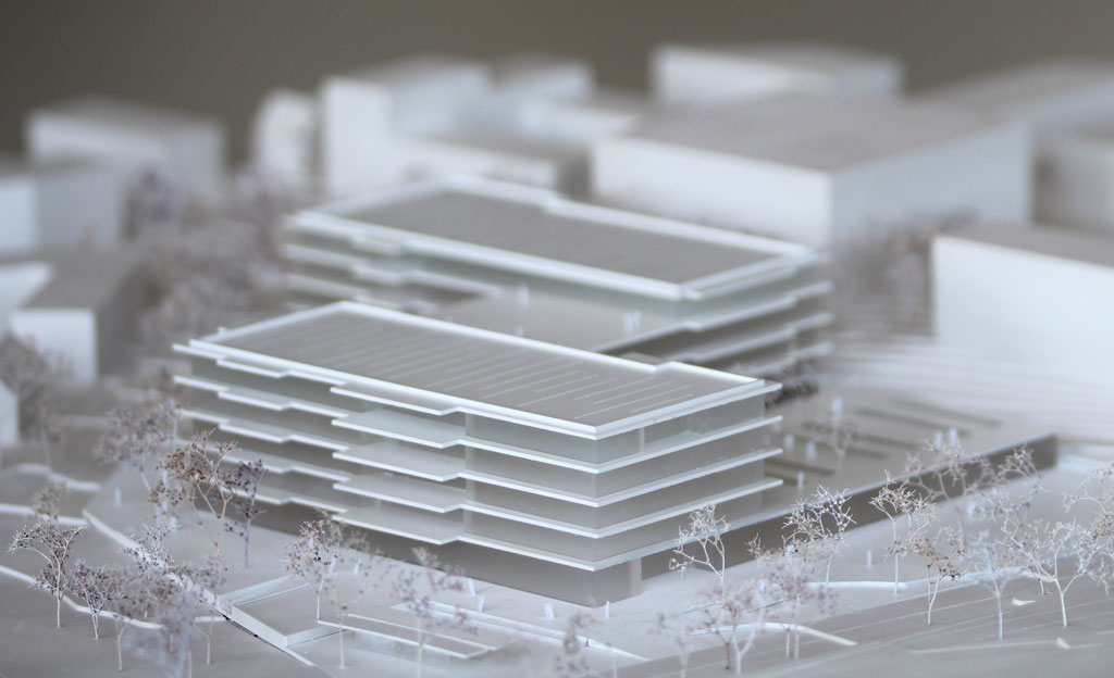 Foto von einem weißen Architekturmodell mit einfachen Kuben für die Umgebungsgebäude und dem detaillierter ausgeführten Schulneubau. Dieser besteht aus zwei parallelen länglichen Riegeln, die versetzt zu einander angeordnet sind und die mit einem senkrecht dazu verlaufenden schmäleren Querbau verbunden sind. Die Längsriegel bestehen jeweils aus 5 Geschossen, der Querriegel aus vier Geschossen. Jedes Geschoss ist aus mattiertem Plexiglas gefertigt, die auskragenden Böden und Decken aus weißem Kunststoff. Im Erdgeschoss gibt es eine Sporthalle, deren Dach begehbar und als Aufenthaltsfläche nutzbar ist. Die Bäume sind aus weiß lackierten getrockneten Pflanzen aufgeklebt.
