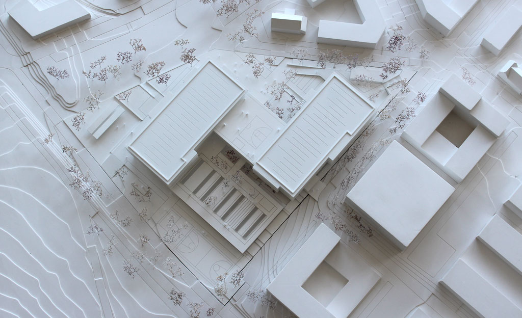 Foto von einem weißen Architekturmodell mit einfachen Kuben für die Umgebungsgebäude und dem detaillierter ausgeführten Schulneubau. Dieser besteht aus zwei parallelen länglichen Riegeln, die versetzt zu einander angeordnet sind und die mit einem senkrecht dazu verlaufenden schmäleren Querbau verbunden sind. Die Längsriegel bestehen jeweils aus 5 Geschossen, der Querriegel aus vier Geschossen. Jedes Geschoss ist aus mattiertem Plexiglas gefertigt, die auskragenden Böden und Decken aus weißem Kunststoff. Im Erdgeschoss gibt es eine Sporthalle, deren Dach begehbar und als Aufenthaltsfläche nutzbar ist. Die Bäume sind aus weiß lackierten getrockneten Pflanzen aufgeklebt.