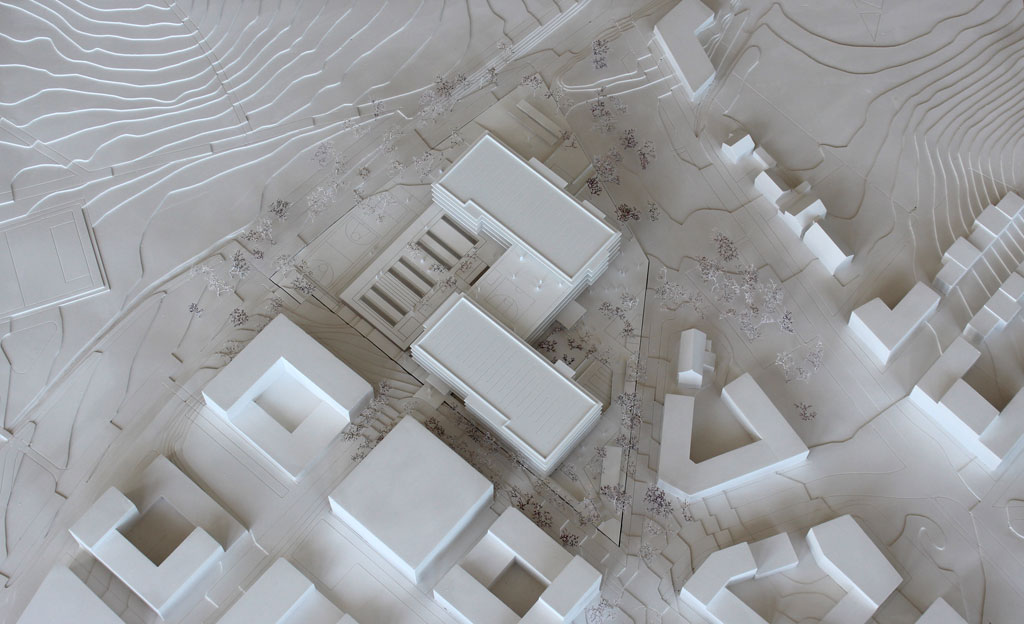 Foto von einem weißen Architekturmodell mit einfachen Kuben für die Umgebungsgebäude und dem detaillierter ausgeführten Schulneubau. Dieser besteht aus zwei parallelen länglichen Riegeln, die versetzt zu einander angeordnet sind und die mit einem senkrecht dazu verlaufenden schmäleren Querbau verbunden sind. Die Längsriegel bestehen jeweils aus 5 Geschossen, der Querriegel aus vier Geschossen. Jedes Geschoss ist aus mattiertem Plexiglas gefertigt, die auskragenden Böden und Decken aus weißem Kunststoff. Im Erdgeschoss gibt es eine Sporthalle, deren Dach begehbar und als Aufenthaltsfläche nutzbar ist. Die Bäume sind aus weiß lackierten getrockneten Pflanzen aufgeklebt.