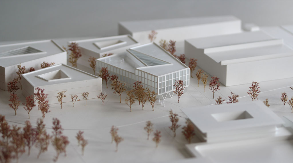 weißes Architekturmodell mit kubischen Umgebungsgebäuden und rötlich gefärbten Bäumen aus getrockneten Pflanzen. Das neu geplante Campusgelände besteht aus einer Anordnung von vier Gebäuden, von denen eines detailliert ausgeführt ist mit weißen Fassadenelementen und dahinterliegenden durchscheinenden Acrylflächen. In der Mitte des Gebäudes liegt ein großes trapezförmiges Atrium, das mit einer Plexiglasfläche abgedeckt ist. Das Erdgeschoss ist durchgehend transparent mit unregelmäig vorgestellten weißen v-förmigen Stützen