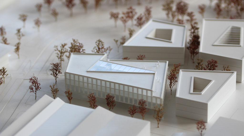 weißes Architekturmodell mit kubischen Umgebungsgebäuden und rötlich gefärbten Bäumen aus getrockneten Pflanzen. Das neu geplante Campusgelände besteht aus einer Anordnung von vier Gebäuden, von denen eines detailliert ausgeführt ist mit weißen Fassadenelementen und dahinterliegenden durchscheinenden Acrylflächen. In der Mitte des Gebäudes liegt ein großes trapezförmiges Atrium, das mit einer Plexiglasfläche abgedeckt ist. Das Erdgeschoss ist durchgehend transparent mit unregelmäig vorgestellten weißen v-förmigen Stützen