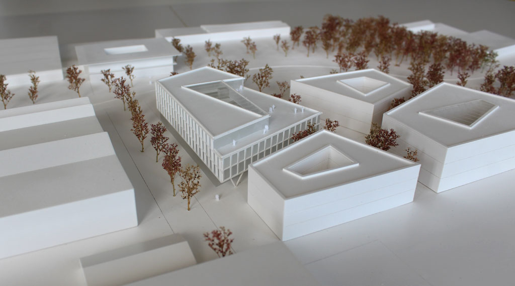 weißes Architekturmodell mit kubischen Umgebungsgebäuden und rötlich gefärbten Bäumen aus getrockneten Pflanzen. Das neu geplante Campusgelände besteht aus einer Anordnung von vier Gebäuden, von denen eines detailliert ausgeführt ist mit weißen Fassadenelementen und dahinterliegenden durchscheinenden Acrylflächen. In der Mitte des Gebäudes liegt ein großes trapezförmiges Atrium, das mit einer Plexiglasfläche abgedeckt ist. Das Erdgeschoss ist durchgehend transparent mit unregelmäig vorgestellten weißen v-förmigen Stützen