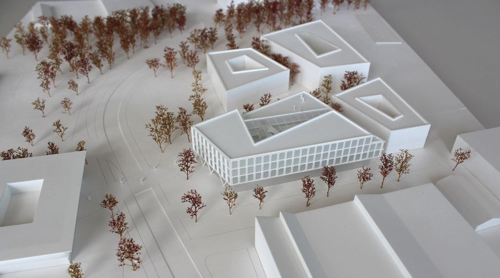 weißes Architekturmodell mit kubischen Umgebungsgebäuden und rötlich gefärbten Bäumen aus getrockneten Pflanzen. Das neu geplante Campusgelände besteht aus einer Anordnung von vier Gebäuden, von denen eines detailliert ausgeführt ist mit weißen Fassadenelementen und dahinterliegenden durchscheinenden Acrylflächen. In der Mitte des Gebäudes liegt ein großes trapezförmiges Atrium, das mit einer Plexiglasfläche abgedeckt ist. Das Erdgeschoss ist durchgehend transparent mit unregelmäig vorgestellten weißen v-förmigen Stützen
