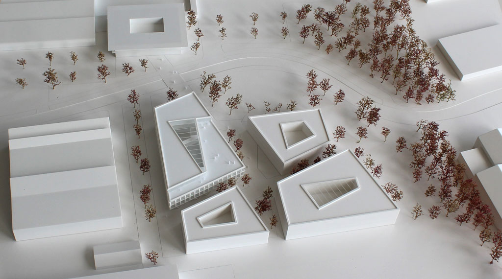 weißes Architekturmodell mit kubischen Umgebungsgebäuden und rötlich gefärbten Bäumen aus getrockneten Pflanzen. Das neu geplante Campusgelände besteht aus einer Anordnung von vier Gebäuden, von denen eines detailliert ausgeführt ist mit weißen Fassadenelementen und dahinterliegenden durchscheinenden Acrylflächen. In der Mitte des Gebäudes liegt ein großes trapezförmiges Atrium, das mit einer Plexiglasfläche abgedeckt ist. Das Erdgeschoss ist durchgehend transparent mit unregelmäig vorgestellten weißen v-förmigen Stützen
