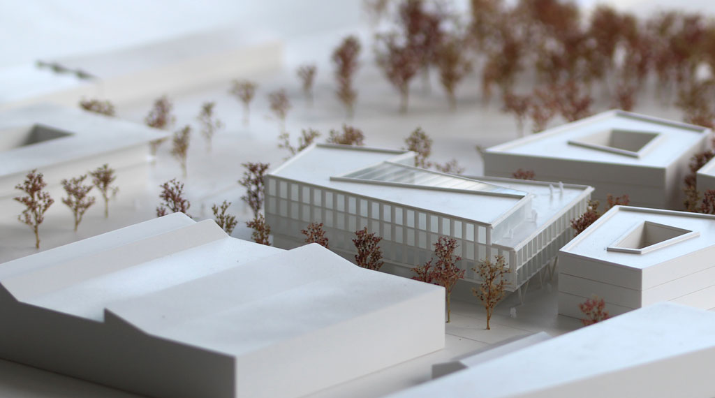 weißes Architekturmodell mit kubischen Umgebungsgebäuden und rötlich gefärbten Bäumen aus getrockneten Pflanzen. Das neu geplante Campusgelände besteht aus einer Anordnung von vier Gebäuden, von denen eines detailliert ausgeführt ist mit weißen Fassadenelementen und dahinterliegenden durchscheinenden Acrylflächen. In der Mitte des Gebäudes liegt ein großes trapezförmiges Atrium, das mit einer Plexiglasfläche abgedeckt ist. Das Erdgeschoss ist durchgehend transparent mit unregelmäig vorgestellten weißen v-förmigen Stützen