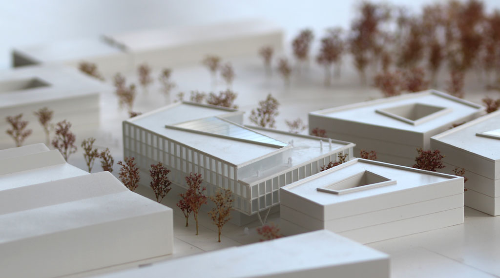 weißes Architekturmodell mit kubischen Umgebungsgebäuden und rötlich gefärbten Bäumen aus getrockneten Pflanzen. Das neu geplante Campusgelände besteht aus einer Anordnung von vier Gebäuden, von denen eines detailliert ausgeführt ist mit weißen Fassadenelementen und dahinterliegenden durchscheinenden Acrylflächen. In der Mitte des Gebäudes liegt ein großes trapezförmiges Atrium, das mit einer Plexiglasfläche abgedeckt ist. Das Erdgeschoss ist durchgehend transparent mit unregelmäig vorgestellten weißen v-förmigen Stützen
