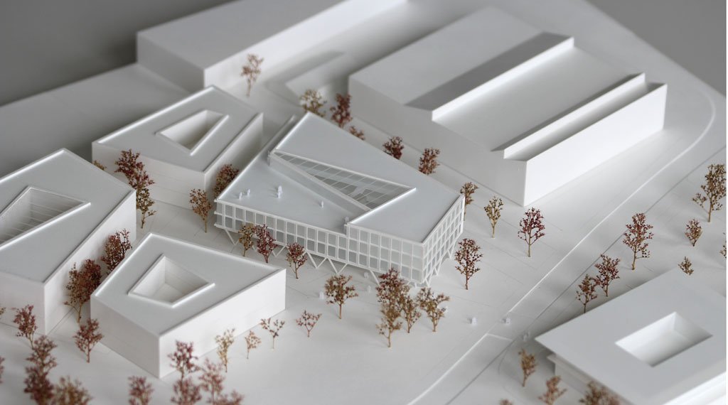 weißes Architekturmodell mit kubischen Umgebungsgebäuden und rötlich gefärbten Bäumen aus getrockneten Pflanzen. Das neu geplante Campusgelände besteht aus einer Anordnung von vier Gebäuden, von denen eines detailliert ausgeführt ist mit weißen Fassadenelementen und dahinterliegenden durchscheinenden Acrylflächen. In der Mitte des Gebäudes liegt ein großes trapezförmiges Atrium, das mit einer Plexiglasfläche abgedeckt ist. Das Erdgeschoss ist durchgehend transparent mit unregelmäig vorgestellten weißen v-förmigen Stützen