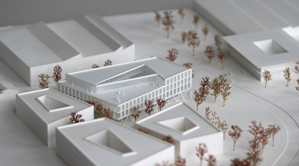 weißes Architekturmodell mit kubischen Umgebungsgebäuden und rötlich gefärbten Bäumen aus getrockneten Pflanzen. Das neu geplante Campusgelände besteht aus einer Anordnung von vier Gebäuden, von denen eines detailliert ausgeführt ist mit weißen Fassadenelementen und dahinterliegenden durchscheinenden Acrylflächen. In der Mitte des Gebäudes liegt ein großes trapezförmiges Atrium, das mit einer Plexiglasfläche abgedeckt ist. Das Erdgeschoss ist durchgehend transparent mit unregelmäig vorgestellten weißen v-förmigen Stützen