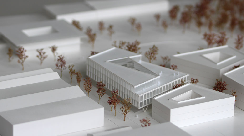 weißes Architekturmodell mit kubischen Umgebungsgebäuden und rötlich gefärbten Bäumen aus getrockneten Pflanzen. Das neu geplante Campusgelände besteht aus einer Anordnung von vier Gebäuden, von denen eines detailliert ausgeführt ist mit weißen Fassadenelementen und dahinterliegenden durchscheinenden Acrylflächen. In der Mitte des Gebäudes liegt ein großes trapezförmiges Atrium, das mit einer Plexiglasfläche abgedeckt ist. Das Erdgeschoss ist durchgehend transparent mit unregelmäig vorgestellten weißen v-förmigen Stützen