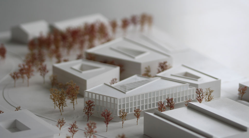 weißes Architekturmodell mit kubischen Umgebungsgebäuden und rötlich gefärbten Bäumen aus getrockneten Pflanzen. Das neu geplante Campusgelände besteht aus einer Anordnung von vier Gebäuden, von denen eines detailliert ausgeführt ist mit weißen Fassadenelementen und dahinterliegenden durchscheinenden Acrylflächen. In der Mitte des Gebäudes liegt ein großes trapezförmiges Atrium, das mit einer Plexiglasfläche abgedeckt ist. Das Erdgeschoss ist durchgehend transparent mit unregelmäig vorgestellten weißen v-förmigen Stützen
