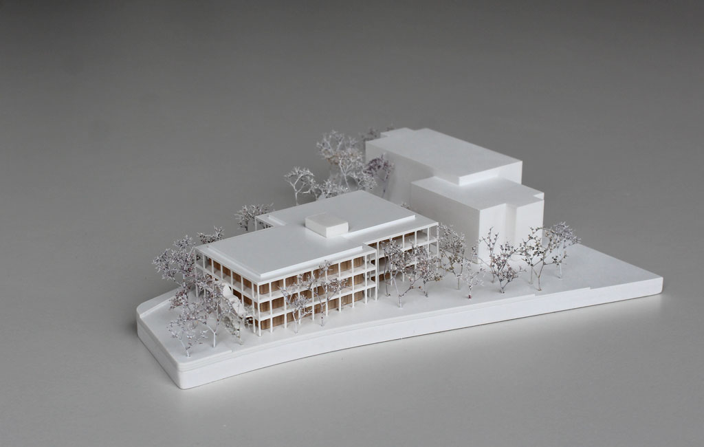 weißes Architekturmodell mit einfachem kubenförmigem Bestandsbau und detaillierterem Neubau. Dieser ist aus Schichten aufgebaut, weiße Böden und Decken, die Geschosse dazwischen aus Holz. Es gibt auskragende Balkone rundherum, die mit dünnen weißen Stützen begrenzt sind. Die Bäume auf dem Grundstücke sind aus weiß lackierten getrockneten Pflanzen.