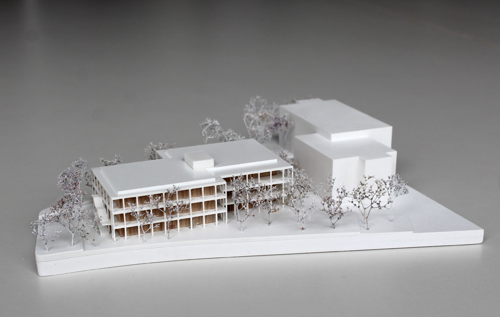 weißes Architekturmodell mit einfachem kubenförmigem Bestandsbau und detaillierterem Neubau. Dieser ist aus Schichten aufgebaut, weiße Böden und Decken, die Geschosse dazwischen aus Holz. Es gibt auskragende Balkone rundherum, die mit dünnen weißen Stützen begrenzt sind. Die Bäume auf dem Grundstücke sind aus weiß lackierten getrockneten Pflanzen.