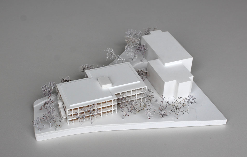 weißes Architekturmodell mit einfachem kubenförmigem Bestandsbau und detaillierterem Neubau. Dieser ist aus Schichten aufgebaut, weiße Böden und Decken, die Geschosse dazwischen aus Holz. Es gibt auskragende Balkone rundherum, die mit dünnen weißen Stützen begrenzt sind. Die Bäume auf dem Grundstücke sind aus weiß lackierten getrockneten Pflanzen.