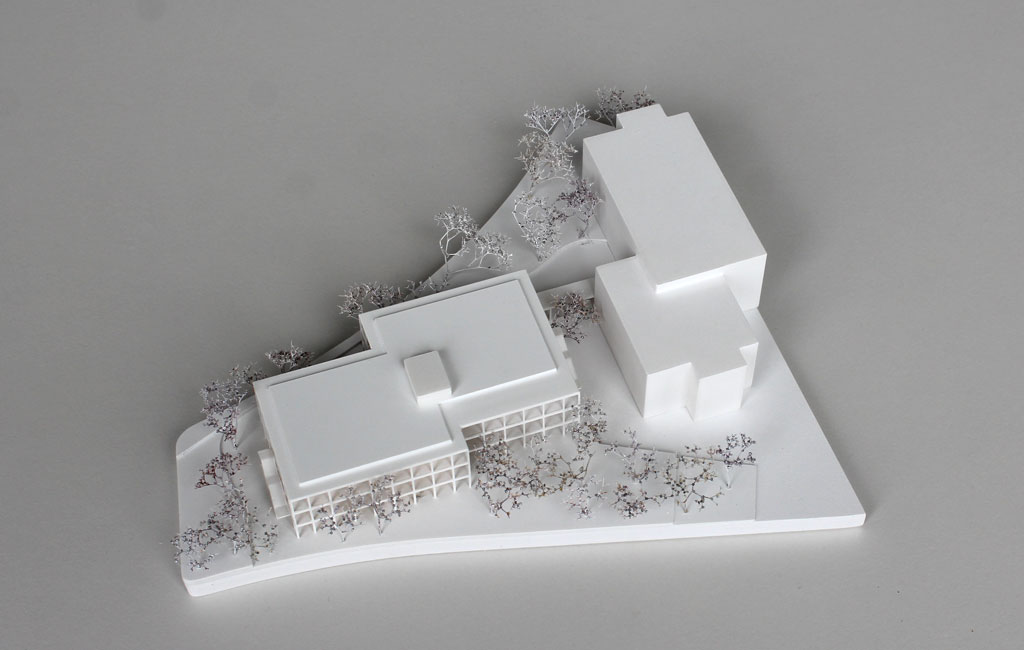 weißes Architekturmodell mit einfachem kubenförmigem Bestandsbau und detaillierterem Neubau. Dieser ist aus Schichten aufgebaut, weiße Böden und Decken, die Geschosse dazwischen aus Holz. Es gibt auskragende Balkone rundherum, die mit dünnen weißen Stützen begrenzt sind. Die Bäume auf dem Grundstücke sind aus weiß lackierten getrockneten Pflanzen.