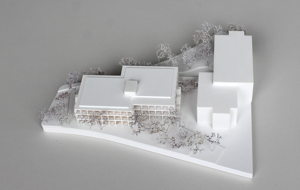 weißes Architekturmodell mit einfachem kubenförmigem Bestandsbau und detaillierterem Neubau. Dieser ist aus Schichten aufgebaut, weiße Böden und Decken, die Geschosse dazwischen aus Holz. Es gibt auskragende Balkone rundherum, die mit dünnen weißen Stützen begrenzt sind. Die Bäume auf dem Grundstücke sind aus weiß lackierten getrockneten Pflanzen.