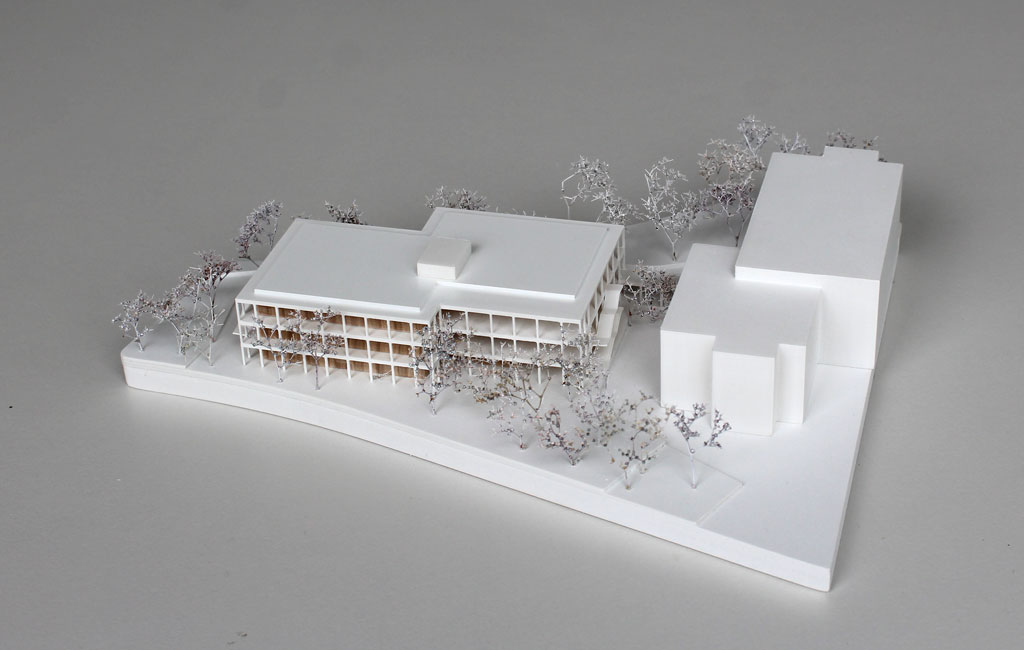 weißes Architekturmodell mit einfachem kubenförmigem Bestandsbau und detaillierterem Neubau. Dieser ist aus Schichten aufgebaut, weiße Böden und Decken, die Geschosse dazwischen aus Holz. Es gibt auskragende Balkone rundherum, die mit dünnen weißen Stützen begrenzt sind. Die Bäume auf dem Grundstücke sind aus weiß lackierten getrockneten Pflanzen.