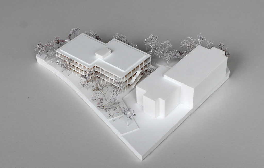 weißes Architekturmodell mit einfachem kubenförmigem Bestandsbau und detaillierterem Neubau. Dieser ist aus Schichten aufgebaut, weiße Böden und Decken, die Geschosse dazwischen aus Holz. Es gibt auskragende Balkone rundherum, die mit dünnen weißen Stützen begrenzt sind. Die Bäume auf dem Grundstücke sind aus weiß lackierten getrockneten Pflanzen.