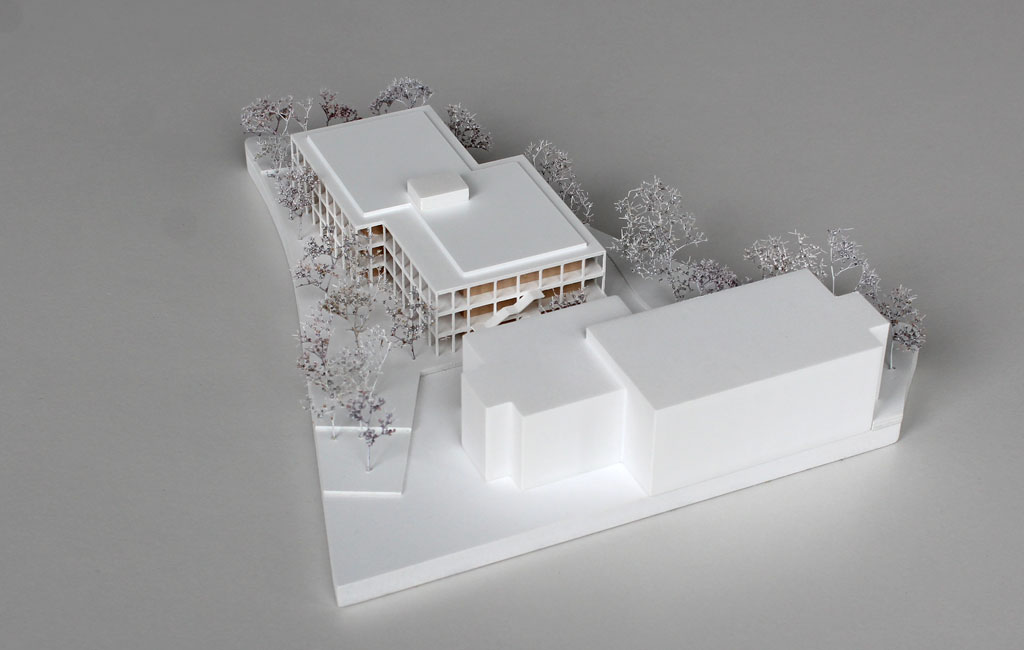 weißes Architekturmodell mit einfachem kubenförmigem Bestandsbau und detaillierterem Neubau. Dieser ist aus Schichten aufgebaut, weiße Böden und Decken, die Geschosse dazwischen aus Holz. Es gibt auskragende Balkone rundherum, die mit dünnen weißen Stützen begrenzt sind. Die Bäume auf dem Grundstücke sind aus weiß lackierten getrockneten Pflanzen.