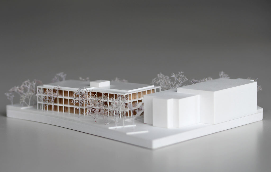 weißes Architekturmodell mit einfachem kubenförmigem Bestandsbau und detaillierterem Neubau. Dieser ist aus Schichten aufgebaut, weiße Böden und Decken, die Geschosse dazwischen aus Holz. Es gibt auskragende Balkone rundherum, die mit dünnen weißen Stützen begrenzt sind. Die Bäume auf dem Grundstücke sind aus weiß lackierten getrockneten Pflanzen.