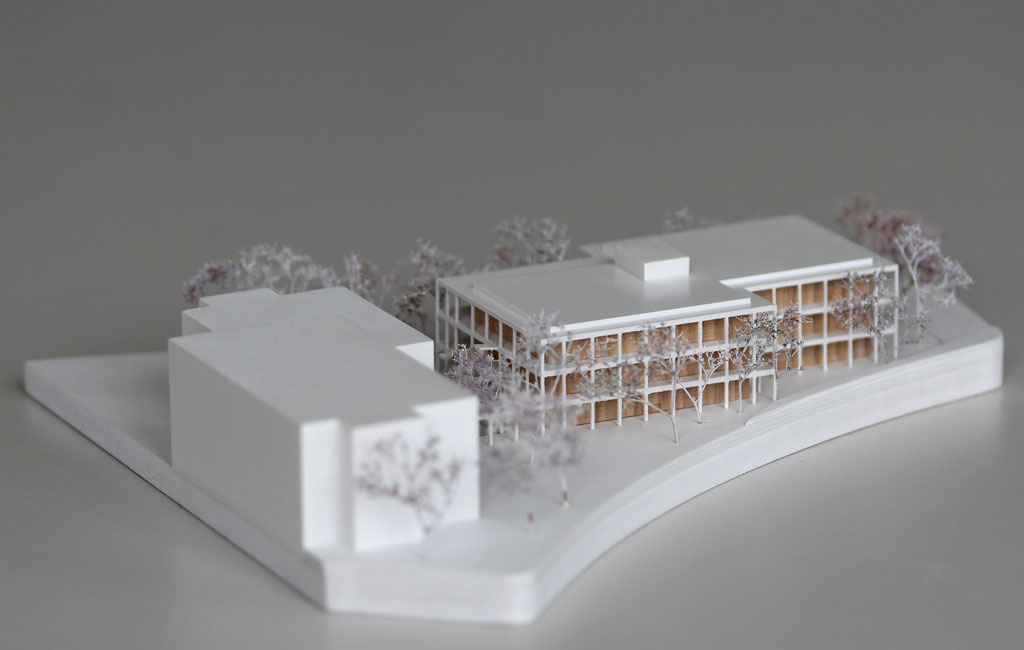 weißes Architekturmodell mit einfachem kubenförmigem Bestandsbau und detaillierterem Neubau. Dieser ist aus Schichten aufgebaut, weiße Böden und Decken, die Geschosse dazwischen aus Holz. Es gibt auskragende Balkone rundherum, die mit dünnen weißen Stützen begrenzt sind. Die Bäume auf dem Grundstücke sind aus weiß lackierten getrockneten Pflanzen.