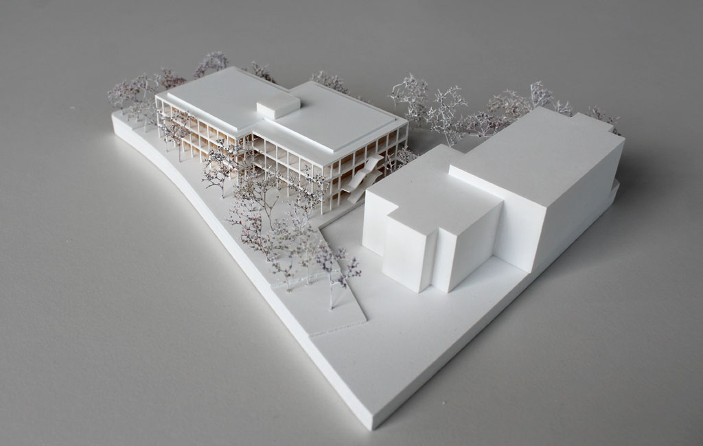 weißes Architekturmodell mit einfachem kubenförmigem Bestandsbau und detaillierterem Neubau. Dieser ist aus Schichten aufgebaut, weiße Böden und Decken, die Geschosse dazwischen aus Holz. Es gibt auskragende Balkone rundherum, die mit dünnen weißen Stützen begrenzt sind. Die Bäume auf dem Grundstücke sind aus weiß lackierten getrockneten Pflanzen.