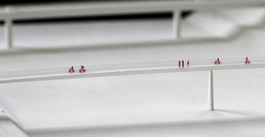 weißes Modell einer schmalen Brück über den Rhein. von seitlich fotografiert. Nahaufnahme der Maßstabfiguren auf dem Rad und zu Fuß aus rotem Acryl