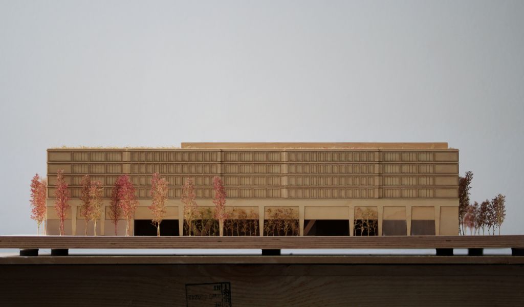 Architekturmodell aus Holz von einem 6-geschossigen Gebäude. Auf dem zweigeschossigen durchlaufenden Sockel gibt es einen L-förmigen Baukörper mit vier Geschossen, sowie ein rechteckiger Baukörper für die Sporthalle. Dazwischen bleibt ein Freibereich, der bepflanzt ist. Auf der Gebäuderückseite gibt es eine aufgeständerten Freibereich, der auch bepflanzt ist.