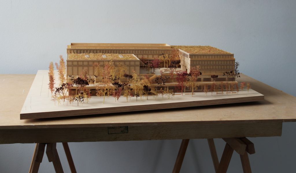 Architekturmodell aus Holz von einem 6-geschossigen Gebäude. Auf dem zweigeschossigen durchlaufenden Sockel gibt es einen L-förmigen Baukörper mit vier Geschossen, sowie ein rechteckiger Baukörper für die Sporthalle. Dazwischen bleibt ein Freibereich, der bepflanzt ist. Auf der Gebäuderückseite gibt es eine aufgeständerten Freibereich, der auch bepflanzt ist.