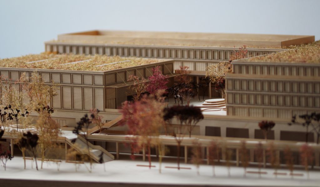 Architekturmodell aus Holz von einem 6-geschossigen Gebäude. Auf dem zweigeschossigen durchlaufenden Sockel gibt es einen L-förmigen Baukörper mit vier Geschossen, sowie ein rechteckiger Baukörper für die Sporthalle. Dazwischen bleibt ein Freibereich, der bepflanzt ist. Auf der Gebäuderückseite gibt es eine aufgeständerten Freibereich, der auch bepflanzt ist.