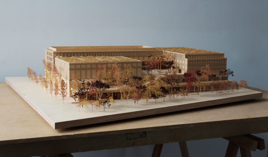 Architekturmodell aus Holz von einem 6-geschossigen Gebäude. Auf dem zweigeschossigen durchlaufenden Sockel gibt es einen L-förmigen Baukörper mit vier Geschossen, sowie ein rechteckiger Baukörper für die Sporthalle. Dazwischen bleibt ein Freibereich, der bepflanzt ist. Auf der Gebäuderückseite gibt es eine aufgeständerten Freibereich, der auch bepflanzt ist.