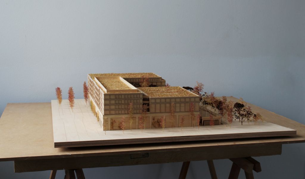 Architekturmodell aus Holz von einem 6-geschossigen Gebäude. Auf dem zweigeschossigen durchlaufenden Sockel gibt es einen L-förmigen Baukörper mit vier Geschossen, sowie ein rechteckiger Baukörper für die Sporthalle. Dazwischen bleibt ein Freibereich, der bepflanzt ist. Auf der Gebäuderückseite gibt es eine aufgeständerten Freibereich, der auch bepflanzt ist.
