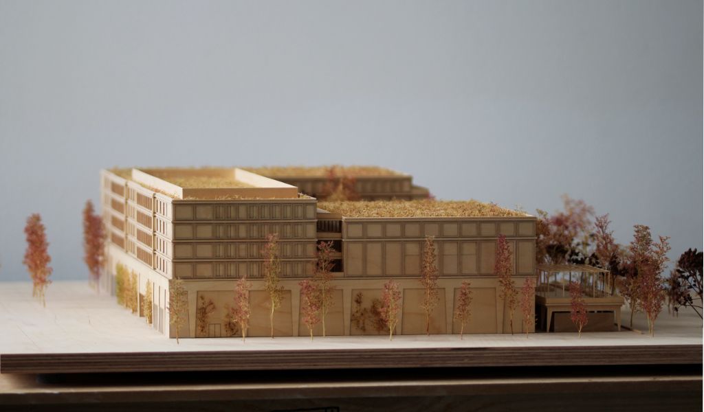 Architekturmodell aus Holz von einem 6-geschossigen Gebäude. Auf dem zweigeschossigen durchlaufenden Sockel gibt es einen L-förmigen Baukörper mit vier Geschossen, sowie ein rechteckiger Baukörper für die Sporthalle. Dazwischen bleibt ein Freibereich, der bepflanzt ist. Auf der Gebäuderückseite gibt es eine aufgeständerten Freibereich, der auch bepflanzt ist.