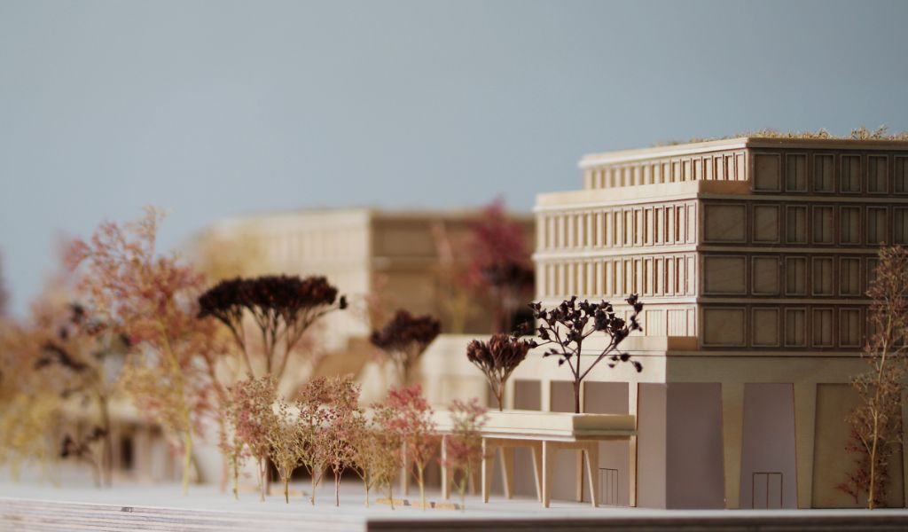 Architekturmodell aus Holz von einem 6-geschossigen Gebäude. Auf dem zweigeschossigen durchlaufenden Sockel gibt es einen L-förmigen Baukörper mit vier Geschossen, sowie ein rechteckiger Baukörper für die Sporthalle. Dazwischen bleibt ein Freibereich, der bepflanzt ist. Auf der Gebäuderückseite gibt es eine aufgeständerten Freibereich, der auch bepflanzt ist.