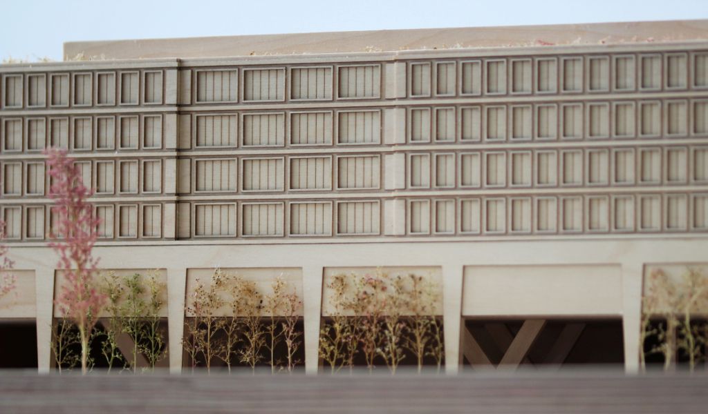 Architekturmodell aus Holz von einem 6-geschossigen Gebäude. Auf dem zweigeschossigen durchlaufenden Sockel gibt es einen L-förmigen Baukörper mit vier Geschossen, sowie ein rechteckiger Baukörper für die Sporthalle. Dazwischen bleibt ein Freibereich, der bepflanzt ist. Auf der Gebäuderückseite gibt es eine aufgeständerten Freibereich, der auch bepflanzt ist.