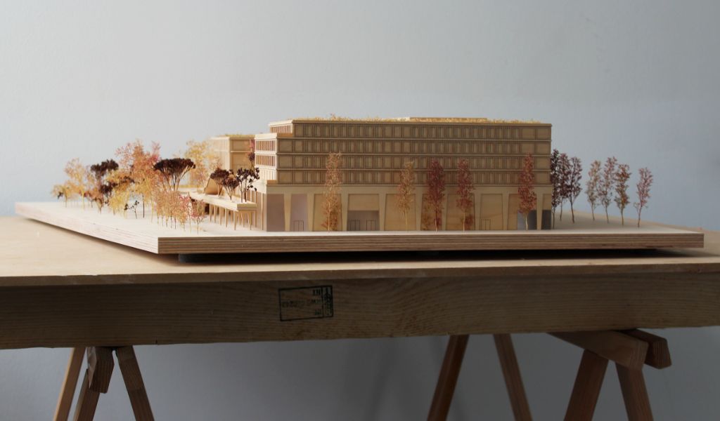 Architekturmodell aus Holz von einem 6-geschossigen Gebäude. Auf dem zweigeschossigen durchlaufenden Sockel gibt es einen L-förmigen Baukörper mit vier Geschossen, sowie ein rechteckiger Baukörper für die Sporthalle. Dazwischen bleibt ein Freibereich, der bepflanzt ist. Auf der Gebäuderückseite gibt es eine aufgeständerten Freibereich, der auch bepflanzt ist.