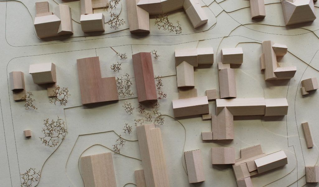 Architekturmodell mit Umgebungsgebäuden aus Lindenholz und zwei Neubauten aus rötlichem Elsbeerenholz. Um die Neubauten herum stehen Bäume aus getrockneten Pflanzen. Die Höhenschichten sind aus gelblicher Finnpappe mit dünnen dunkelbraunen Gravurlinien für die Strassenkanten.