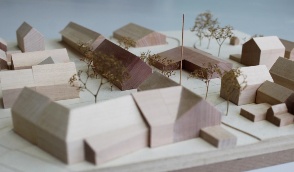 Architekturmodell mit Umgebungsgebäuden aus Lindenholz und zwei Neubauten aus rötlichem Elsbeerenholz. Um die Neubauten herum stehen Bäume aus getrockneten Pflanzen. Die Höhenschichten sind aus gelblicher Finnpappe mit dünnen dunkelbraunen Gravurlinien für die Strassenkanten.