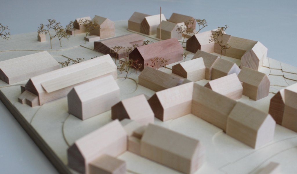 Architekturmodell mit Umgebungsgebäuden aus Lindenholz und zwei Neubauten aus rötlichem Elsbeerenholz. Um die Neubauten herum stehen Bäume aus getrockneten Pflanzen. Die Höhenschichten sind aus gelblicher Finnpappe mit dünnen dunkelbraunen Gravurlinien für die Strassenkanten.