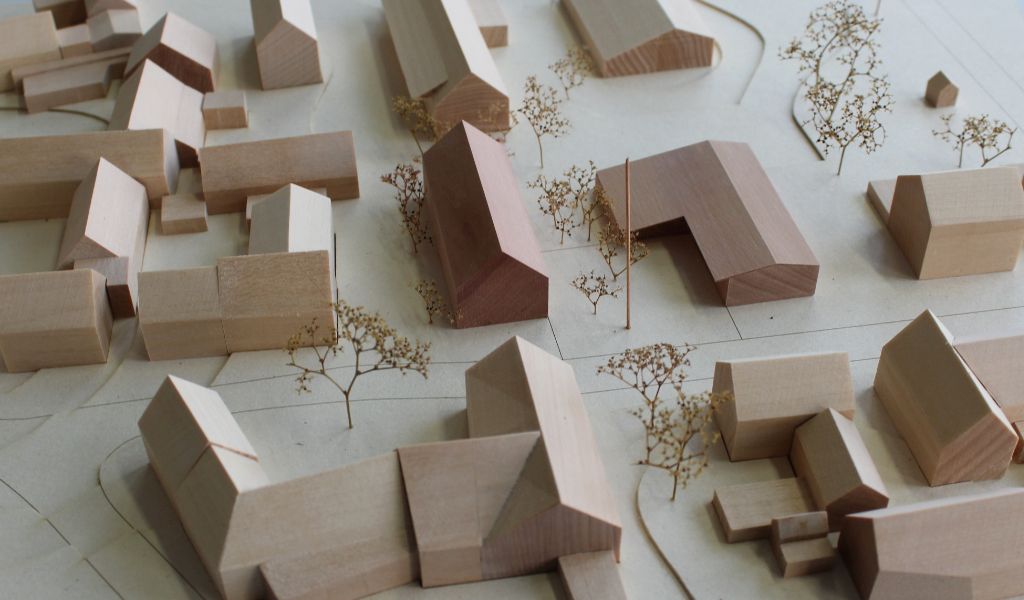 Architekturmodell mit Umgebungsgebäuden aus Lindenholz und zwei Neubauten aus rötlichem Elsbeerenholz. Um die Neubauten herum stehen Bäume aus getrockneten Pflanzen. Die Höhenschichten sind aus gelblicher Finnpappe mit dünnen dunkelbraunen Gravurlinien für die Strassenkanten.