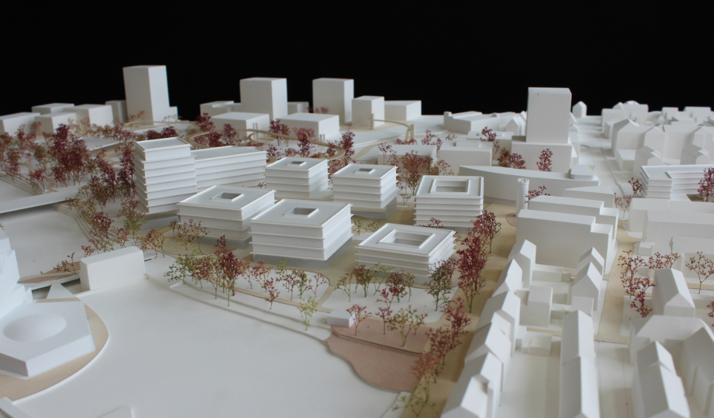weißes Städtebau-Architekturmodell mit frabig angelegten Freiflächen Wegen. Die Bestandsgebäude sind einfache weiße Kuben, die neugeplanten Gebäude haben jeweils einen tranparentes Sockelgeschoss, die weißen Geschossdecken sind jeweils leicht auskragend, die oberen Geschosse sind jeweils weiß. Die Bäume sind rötlich aus getrockneten Pflanzen. Als Wegeverbindung zwischen den bereits bestehenden Campi und der neuen Anlage gibt es einen aufgeständerten 'Loop', ein elliptischer Fuß- und Radweg, der im Modell aus dünnem Holz dargestellt ist.