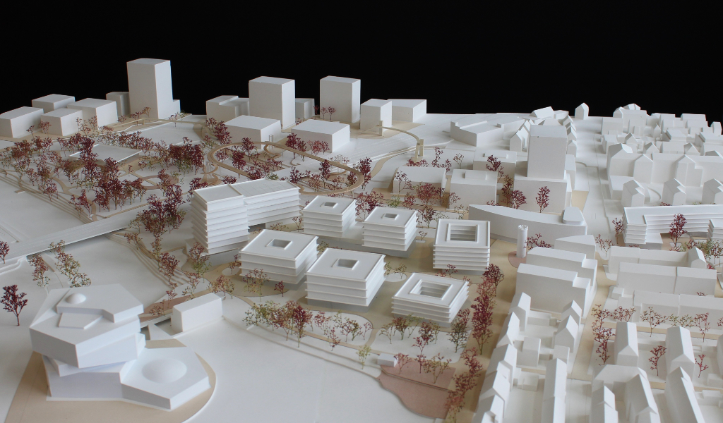 weißes Städtebau-Architekturmodell mit frabig angelegten Freiflächen Wegen. Die Bestandsgebäude sind einfache weiße Kuben, die neugeplanten Gebäude haben jeweils einen tranparentes Sockelgeschoss, die weißen Geschossdecken sind jeweils leicht auskragend, die oberen Geschosse sind jeweils weiß. Die Bäume sind rötlich aus getrockneten Pflanzen. Als Wegeverbindung zwischen den bereits bestehenden Campi und der neuen Anlage gibt es einen aufgeständerten 'Loop', ein elliptischer Fuß- und Radweg, der im Modell aus dünnem Holz dargestellt ist.