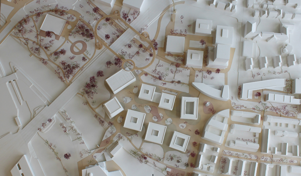 weißes Städtebau-Architekturmodell mit frabig angelegten Freiflächen Wegen. Die Bestandsgebäude sind einfache weiße Kuben, die neugeplanten Gebäude haben jeweils einen tranparentes Sockelgeschoss, die weißen Geschossdecken sind jeweils leicht auskragend, die oberen Geschosse sind jeweils weiß. Die Bäume sind rötlich aus getrockneten Pflanzen. Als Wegeverbindung zwischen den bereits bestehenden Campi und der neuen Anlage gibt es einen aufgeständerten 'Loop', ein elliptischer Fuß- und Radweg, der im Modell aus dünnem Holz dargestellt ist.