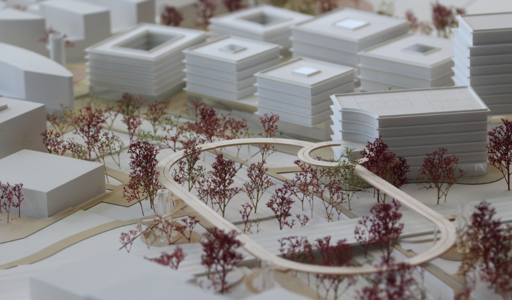 weißes Städtebau-Architekturmodell mit frabig angelegten Freiflächen Wegen. Die Bestandsgebäude sind einfache weiße Kuben, die neugeplanten Gebäude haben jeweils einen tranparentes Sockelgeschoss, die weißen Geschossdecken sind jeweils leicht auskragend, die oberen Geschosse sind jeweils weiß. Die Bäume sind rötlich aus getrockneten Pflanzen. Als Wegeverbindung zwischen den bereits bestehenden Campi und der neuen Anlage gibt es einen aufgeständerten 'Loop', ein elliptischer Fuß- und Radweg, der im Modell aus dünnem Holz dargestellt ist.
