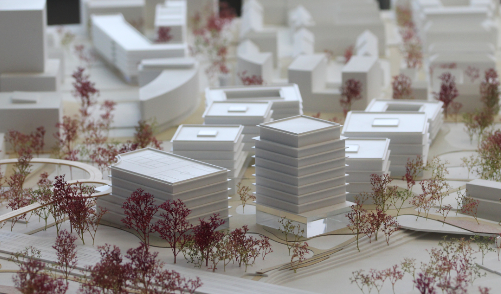 weißes Städtebau-Architekturmodell mit frabig angelegten Freiflächen Wegen. Die Bestandsgebäude sind einfache weiße Kuben, die neugeplanten Gebäude haben jeweils einen tranparentes Sockelgeschoss, die weißen Geschossdecken sind jeweils leicht auskragend, die oberen Geschosse sind jeweils weiß. Die Bäume sind rötlich aus getrockneten Pflanzen. Als Wegeverbindung zwischen den bereits bestehenden Campi und der neuen Anlage gibt es einen aufgeständerten 'Loop', ein elliptischer Fuß- und Radweg, der im Modell aus dünnem Holz dargestellt ist.