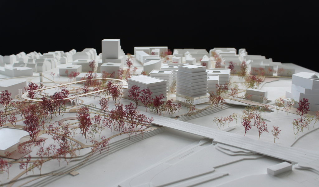 weißes Städtebau-Architekturmodell mit frabig angelegten Freiflächen Wegen. Die Bestandsgebäude sind einfache weiße Kuben, die neugeplanten Gebäude haben jeweils einen tranparentes Sockelgeschoss, die weißen Geschossdecken sind jeweils leicht auskragend, die oberen Geschosse sind jeweils weiß. Die Bäume sind rötlich aus getrockneten Pflanzen. Als Wegeverbindung zwischen den bereits bestehenden Campi und der neuen Anlage gibt es einen aufgeständerten 'Loop', ein elliptischer Fuß- und Radweg, der im Modell aus dünnem Holz dargestellt ist.