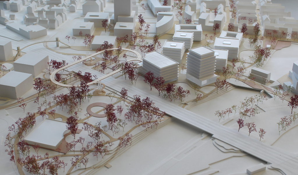 weißes Städtebau-Architekturmodell mit frabig angelegten Freiflächen Wegen. Die Bestandsgebäude sind einfache weiße Kuben, die neugeplanten Gebäude haben jeweils einen tranparentes Sockelgeschoss, die weißen Geschossdecken sind jeweils leicht auskragend, die oberen Geschosse sind jeweils weiß. Die Bäume sind rötlich aus getrockneten Pflanzen. Als Wegeverbindung zwischen den bereits bestehenden Campi und der neuen Anlage gibt es einen aufgeständerten 'Loop', ein elliptischer Fuß- und Radweg, der im Modell aus dünnem Holz dargestellt ist.